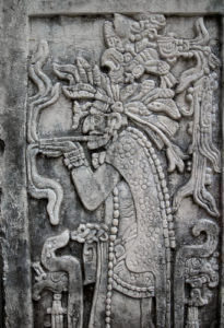 Zusehen ist ein Maya Stein Relief mit einem Maya Priester welcher an einer Pfeife raucht.